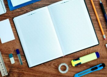 A writing book, pencil, pens, stanley knife, ruler, rubber and other forms of business stationary sitting on a desk