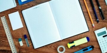 A writing book, pencil, pens, stanley knife, ruler, rubber and other forms of business stationary sitting on a desk