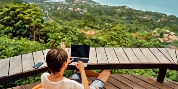 A remote worker sitting in thailand, working from their laptop looking over the forest and beach beyond