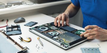 A m,an sitting at a white desk refurbishing a laptop computer with new components