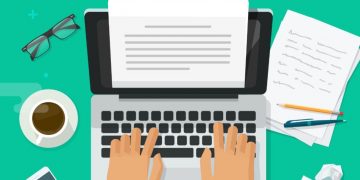 A persons hands writing on a latop with paper, stationary and coffee around