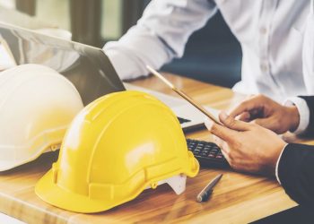 Two colleauges sitting next to each at a desk using health and safety software, with building construction hats symbolising the topic as health and safety