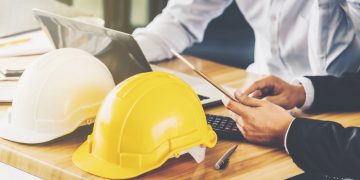 Two colleauges sitting next to each at a desk using health and safety software, with building construction hats symbolising the topic as health and safety