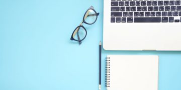 A laptop, reading glasses and notepad sitting on blue surface as an example of the essentials every entrepreneur needs