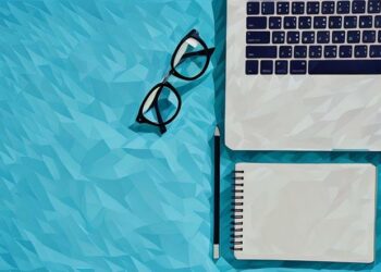 A laptop, reading glasses and notepad sitting on blue surface as an example of the essentials every entrepreneur needs