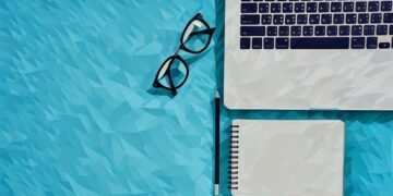 A laptop, reading glasses and notepad sitting on blue surface as an example of the essentials every entrepreneur needs