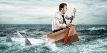 A man using a box to paddle through a shark infested ocean, exampling how good business conituinty planning is enabling him to survive
