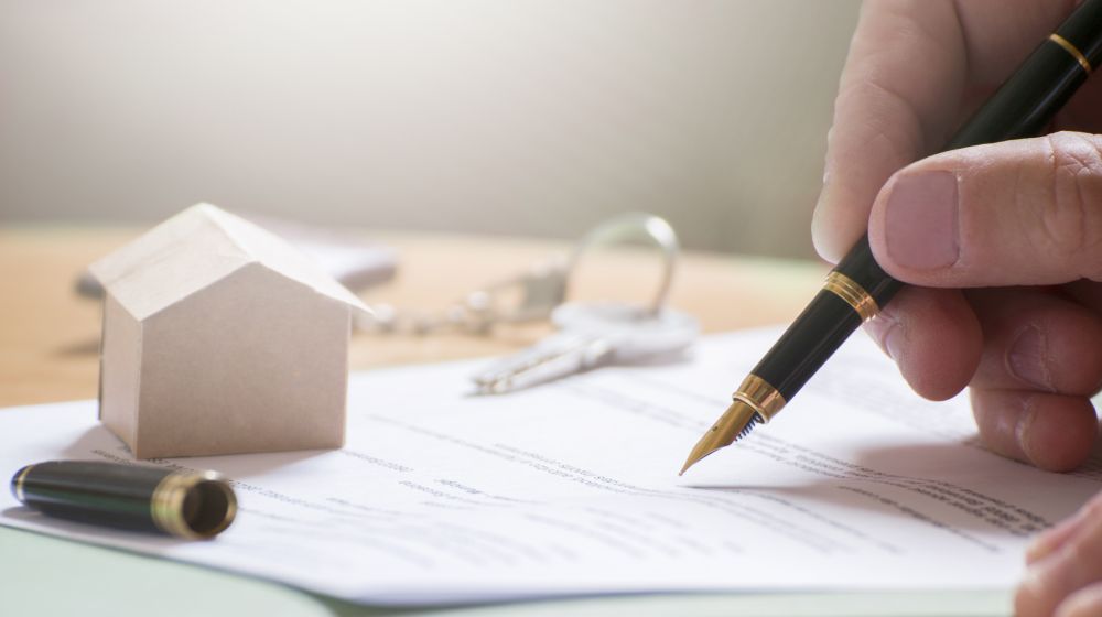 A person signing a mortgage document against the backdrop of a wooden model of the house they've just bought