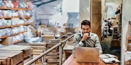 An eCommerce entrepreneur working on the shop, with his product dispatch area in the back filled with packages